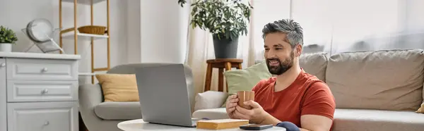 A bearded man in casual attire sits comfortably at home, working remotely on his laptop while enjoying a cup of coffee. — Stock Photo