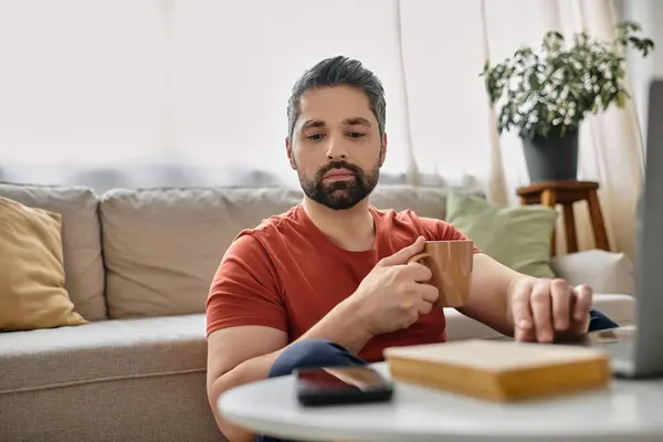 A bearded man in casual attire works remotely on a laptop from home while holding a cup of coffee. — Stock Photo