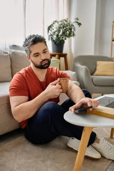 A bearded man in casual attire works remotely from home, sitting on the floor with a coffee mug in one hand and a phone in the other. — Stock Photo