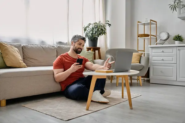 A bearded man sits on the floor in his living room, working on his laptop. He holds a phone in his hand and looks at the screen of the laptop. — Stock Photo