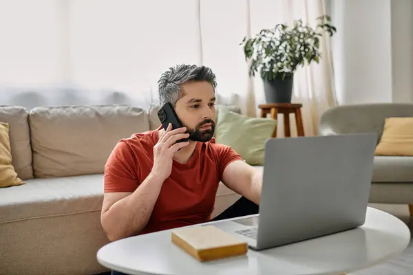 A bearded man in casual attire works remotely from his living room, using a laptop and phone. — Stock Photo