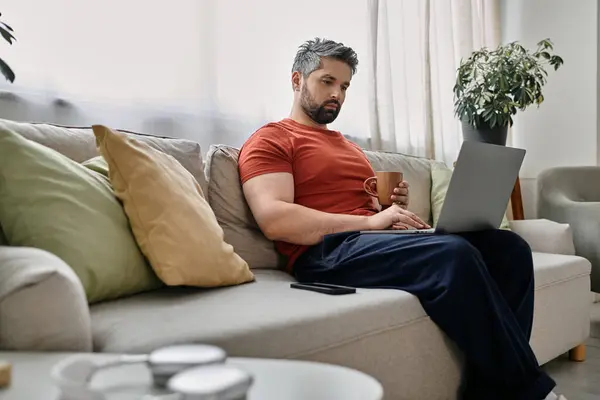 A bearded man in casual attire works remotely from home on a laptop while sitting on a couch and holding a mug. — Stock Photo