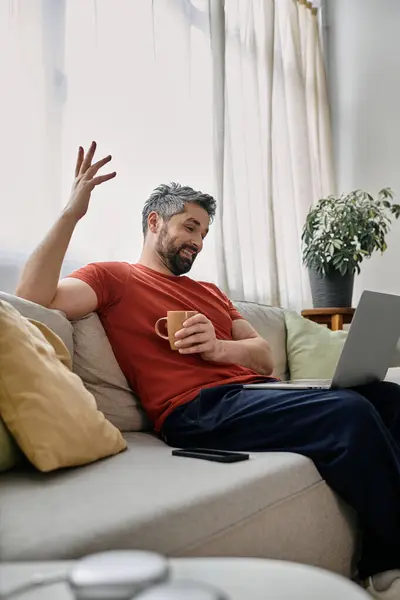 Um homem barbudo vestindo roupas casuais se senta em um sofá em casa, trabalhando em seu laptop enquanto desfruta de uma xícara de café. — Fotografia de Stock