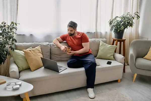 Un hombre barbudo se sienta en un sofá, trabajando en su portátil, rodeado de cómodas almohadas. - foto de stock