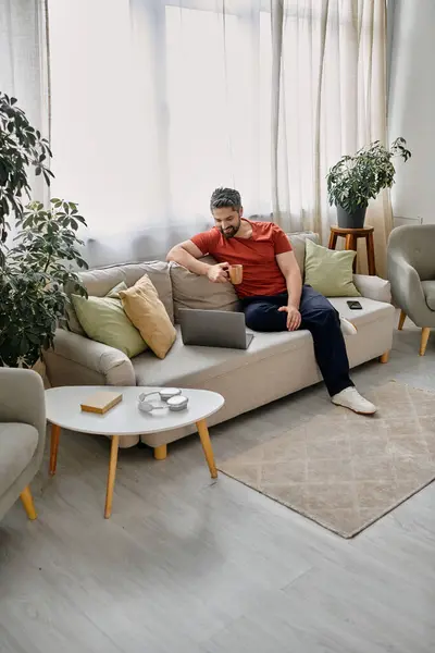 Un uomo barbuto si siede su un divano nella sua casa, lavorando sul suo computer portatile e gustando una tazza di caffè. — Foto stock
