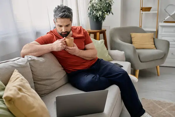 Un uomo barbuto in abbigliamento casual si rilassa su un divano, gustando una tazza di caffè mentre lavora sul suo computer portatile. — Foto stock