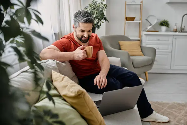 Un hombre barbudo con atuendo casual trabaja remotamente desde casa, sentado en un sofá con una computadora portátil y una taza de café. - foto de stock