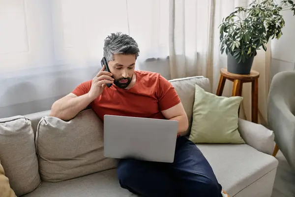 Ein Mann mit Bart, lässig gekleidet, arbeitet von zu Hause aus. Er sitzt auf einer Couch, benutzt einen Laptop und telefoniert mit einem Mobiltelefon. — Stockfoto