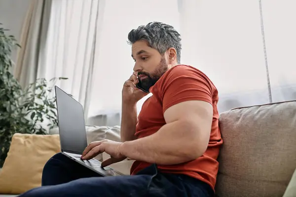 A bearded man in casual clothing works remotely from home, multitasking with his laptop and phone. — Stock Photo