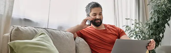 A bearded man in casual attire sits on a couch, working on a laptop and talking on his phone. Natural light streams in through a window behind him. — Stock Photo