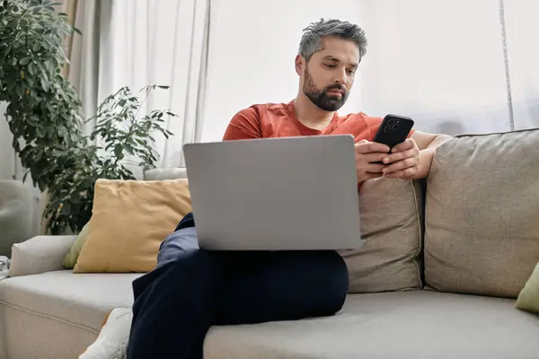 Бородатый мужчина в повседневной одежде работает дистанционно из дома, сидя на диване с ноутбуком и смартфоном. — стоковое фото