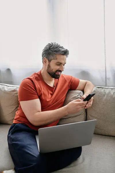 A bearded man sits on a couch in his home, working on a laptop while checking his phone. — Stock Photo