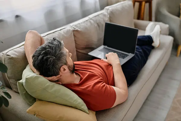 A bearded man works remotely on his laptop while relaxing on a couch at home. — Stock Photo