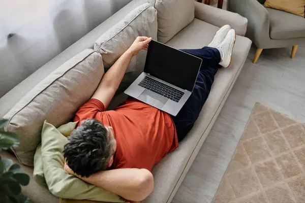 A bearded man in casual attire relaxes on a couch at home, working remotely on his laptop. — Stock Photo