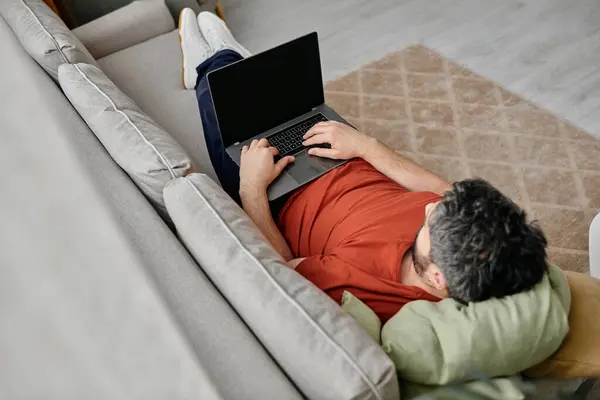 Ein bärtiger Mann liegt mit gestützten Beinen auf einer Couch und arbeitet an einem Laptop. — Stockfoto