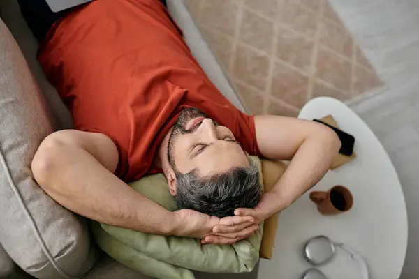 Un hombre barbudo con atuendo casual descansa en un sofá, tomando un descanso de su día de trabajo en casa. - foto de stock