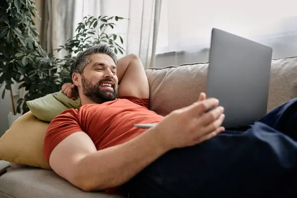 Un uomo barbuto in abbigliamento casual lavora a distanza da casa, comodamente sdraiato su un divano con un computer portatile. — Foto stock