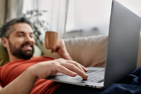 A bearded man wearing casual attire is working remotely from home, comfortably using a laptop while lying on a couch. — Stock Photo
