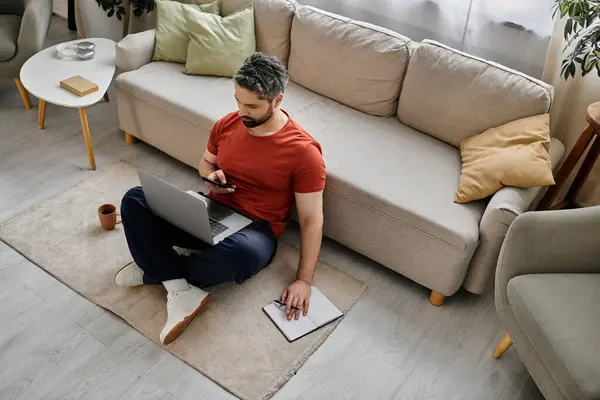 A bearded man sits on the floor, working on his laptop and phone in a casual setting. He is likely working remotely from home. — Stock Photo