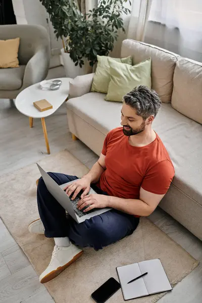 Un uomo barbuto in abbigliamento casual si siede sul pavimento in un ambiente domestico, lavorando su un computer portatile. — Foto stock