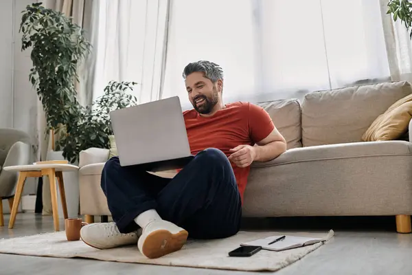 Ein bärtiger Mann sitzt auf einer Couch in seinem Haus und arbeitet an seinem Laptop. — Stockfoto