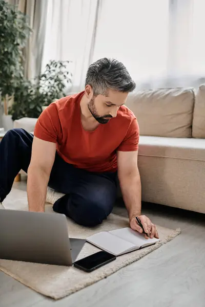 Un homme barbu en tenue décontractée travaille à distance de la maison, assis sur le sol avec un ordinateur portable, un ordinateur portable et un stylo. — Photo de stock