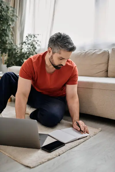 A bearded man in casual attire works remotely from home, sitting on the floor with a laptop, notebook, and pen. — Stock Photo