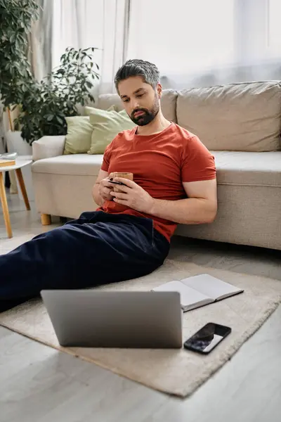 A bearded man in casual attire works remotely from home, sitting on the floor with his laptop and a cup of coffee. — Stock Photo