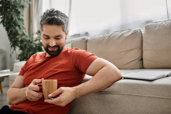 A bearded man in a casual red shirt enjoys a warm drink while working remotely from home. — Stock Photo