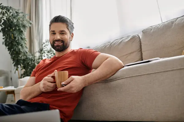 A bearded man in casual attire sits on the floor near a couch, holding a mug, and working remotely from home. — Stock Photo