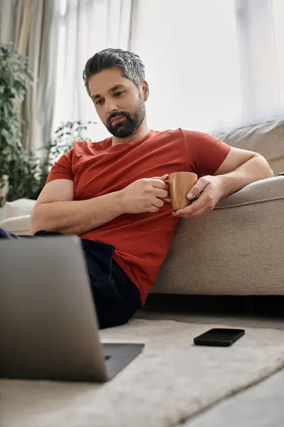 A bearded man in casual attire sits on the floor, working remotely on his laptop with a cup of coffee in his hand. — Stock Photo