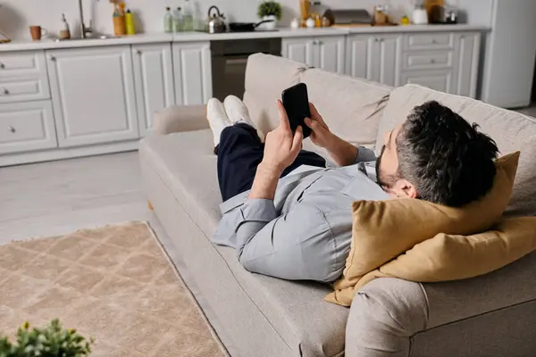 A bearded man in casual attire works remotely from home, lounging on a couch and using a smartphone. — Stock Photo
