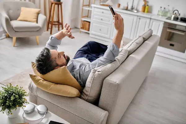 A bearded man in casual attire relaxes on a couch while working remotely on his phone. — Stock Photo