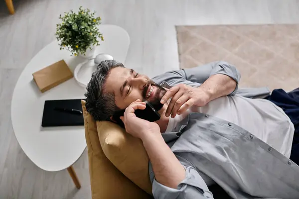 Un homme barbu en tenue décontractée se couche sur un canapé et parle sur son téléphone, profitant d'une pause de son travail à distance. — Photo de stock