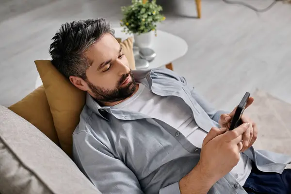 A bearded man in casual attire is lying on a couch, working remotely from home using a smartphone. — Stock Photo