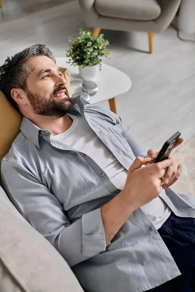 A bearded man in a grey shirt works remotely from home on his couch, holding a smartphone. — Stock Photo