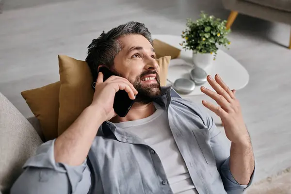 Бородатый мужчина в повседневной одежде лежит на диване, используя свой телефон во время звонка — стоковое фото