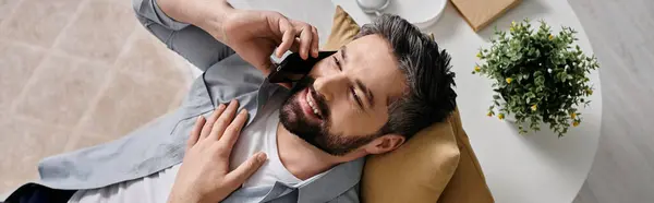Бородатый мужчина в повседневной одежде улыбается, работая удаленно из дома, разговаривая по телефону. — стоковое фото