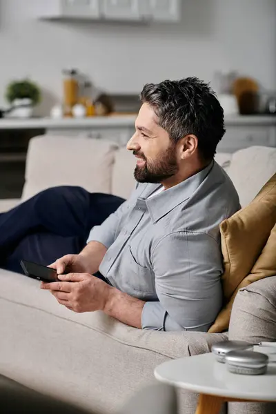 Un hombre barbudo con atuendo casual trabaja en su teléfono mientras se relaja en un sofá. — Stock Photo