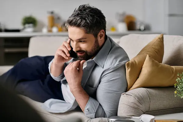 A bearded man in casual attire works remotely from home, making a phone call while lounging on a sofa. — Stock Photo