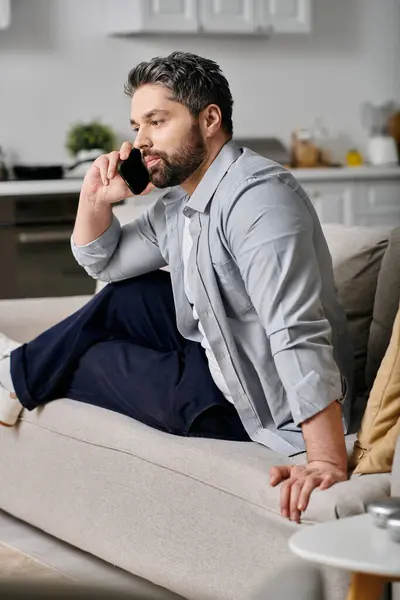 Um homem barbudo de traje casual senta-se em um sofá, trabalhando remotamente em casa. Ele está falando em seu telefone e parece estar focado em seu trabalho. — Fotografia de Stock