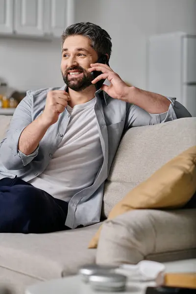A bearded man in a light blue shirt sits on a couch, smiling as he talks on his phone. — Stock Photo