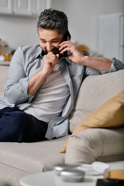 A bearded man in casual attire sits on a couch and talks on the phone while smiling. He appears to be working remotely from home. — Stock Photo