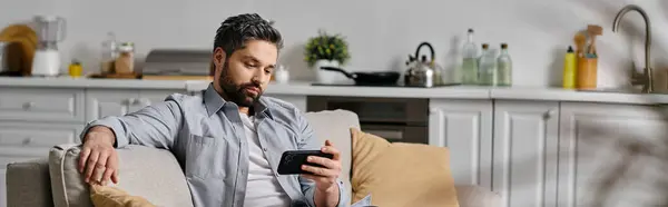 A bearded man in casual attire relaxes on a couch, using his phone in a home setting. — Stock Photo