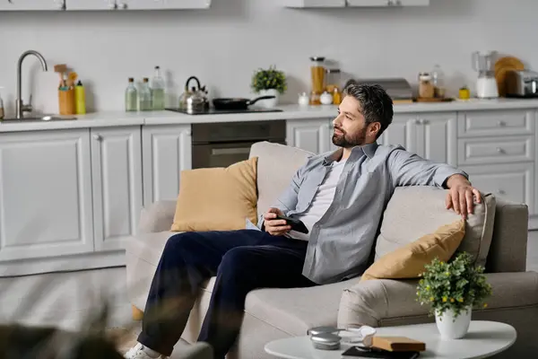 A bearded man in casual attire relaxes on a couch in his home, holding a phone. — Stock Photo