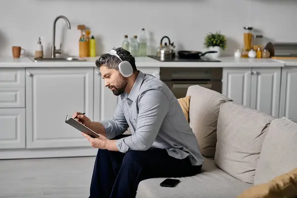 A bearded man wearing headphones sits on a couch in his kitchen — Stock Photo