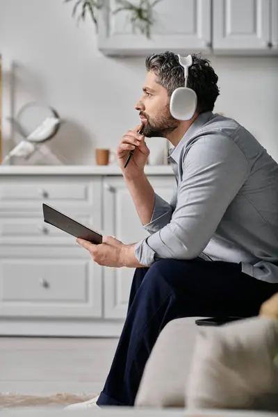 Ein bärtiger Mann sitzt auf einer Couch, hält ein Notizbuch auf dem Schoß und blickt nachdenklich in die Ferne. Er trägt Kopfhörer und ein graues Hemd. — Stockfoto