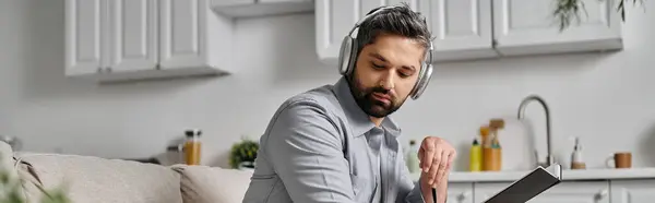 A bearded man in casual attire sitting on a couch in a kitchen, wearing headphones — Stock Photo
