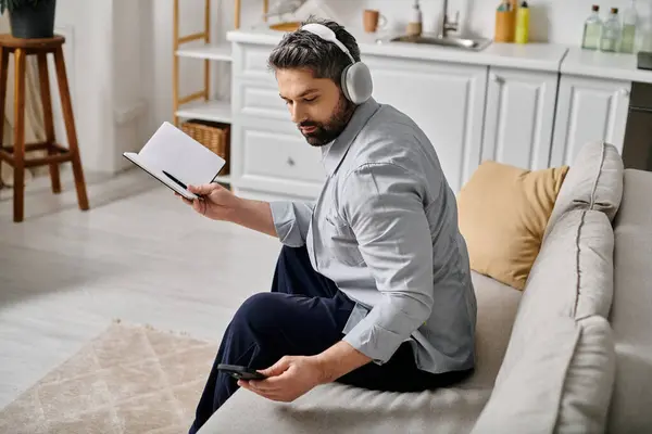 A bearded man in casual attire sits on a couch, working remotely from home. He is using a smartphone and holding a notebook. — Stock Photo