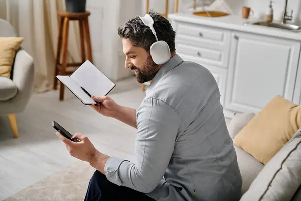 Un hombre barbudo con atuendo casual se sienta en un sofá y trabaja remotamente desde casa. Lleva auriculares y lleva un bloc de notas y un smartphone. - foto de stock